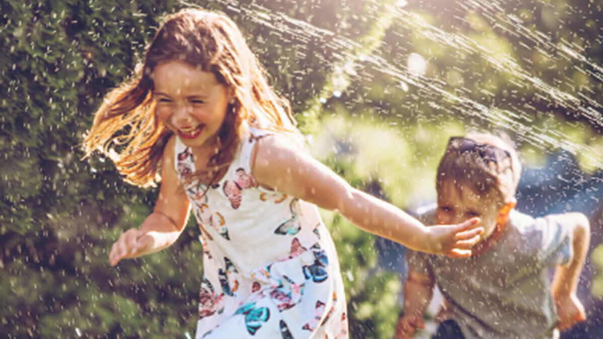 Zwei Kinder, ein Mädchen und ein Junge, spielen fröhlich im Garten und rennen lachend durch einen Wasserstrahl.