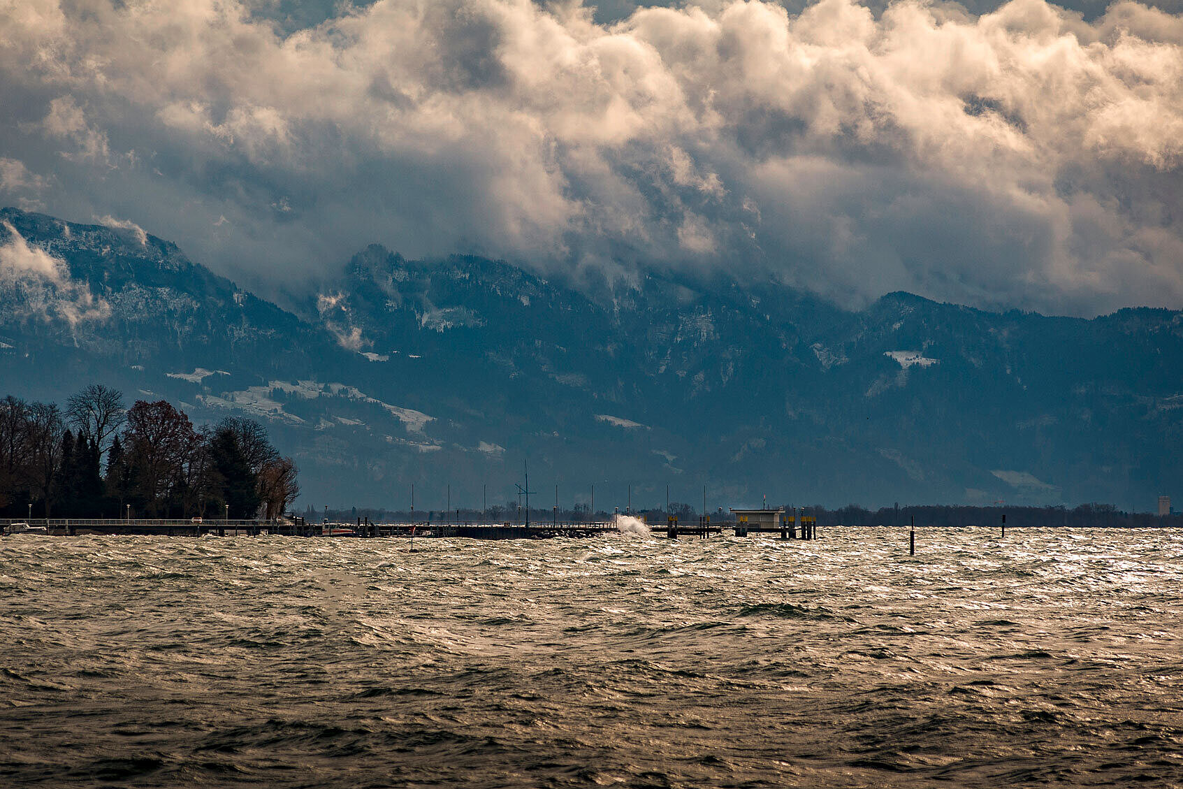 Aufgewühlter Bodensee unter dunklen, dramatisch wirkenden Wolken; im Hintergrund schneebedeckte Berge und eine Uferpromenade mit Bäumen, während der Sturm hohe Wellen erzeugt.