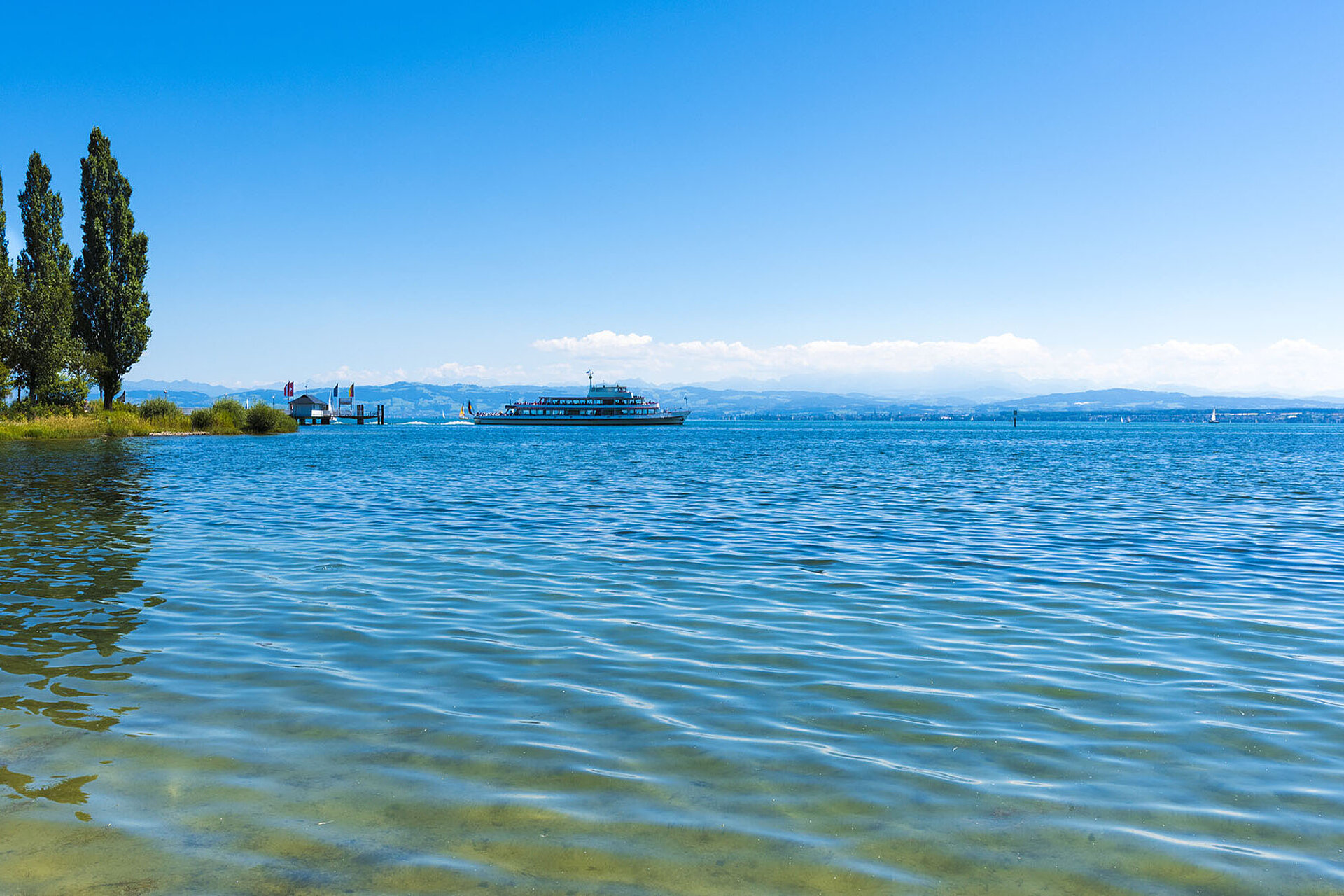 Blick auf den Bodensee an einem sonnigen Tag mit klarem Wasser im Vordergrund, Bäumen am Ufer und einem Ausflugsschiff in der Ferne; im Hintergrund sind die Alpen zu sehen.